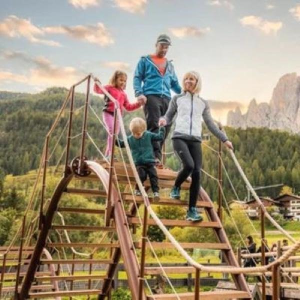 Una famiglia felice attraversa un ponte di legno in un parco giochi, con le montagne della Val di Fassa sullo sfondo