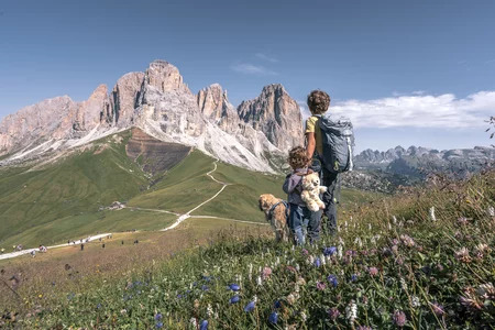 Una famiglia con un bambino e un cane passeggiano lungo un sentiero della Val di Fassa, con un paesaggio estivo mozzafiato sullo sfondo